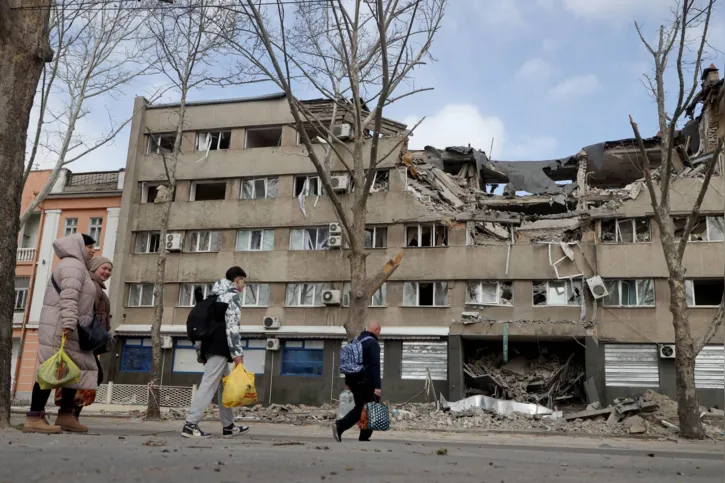 People walk past a damaged building in the Ukrainian city of Mykolaiv on March 27, 2022. - Over the weekend, the air raid sirens were no longer disturbing locals in Mykolaiv, who were increasingly venturing out on the streets. Most barely hurry when they hear them. (Photo by Oleksandr GIMANOV / AFP)