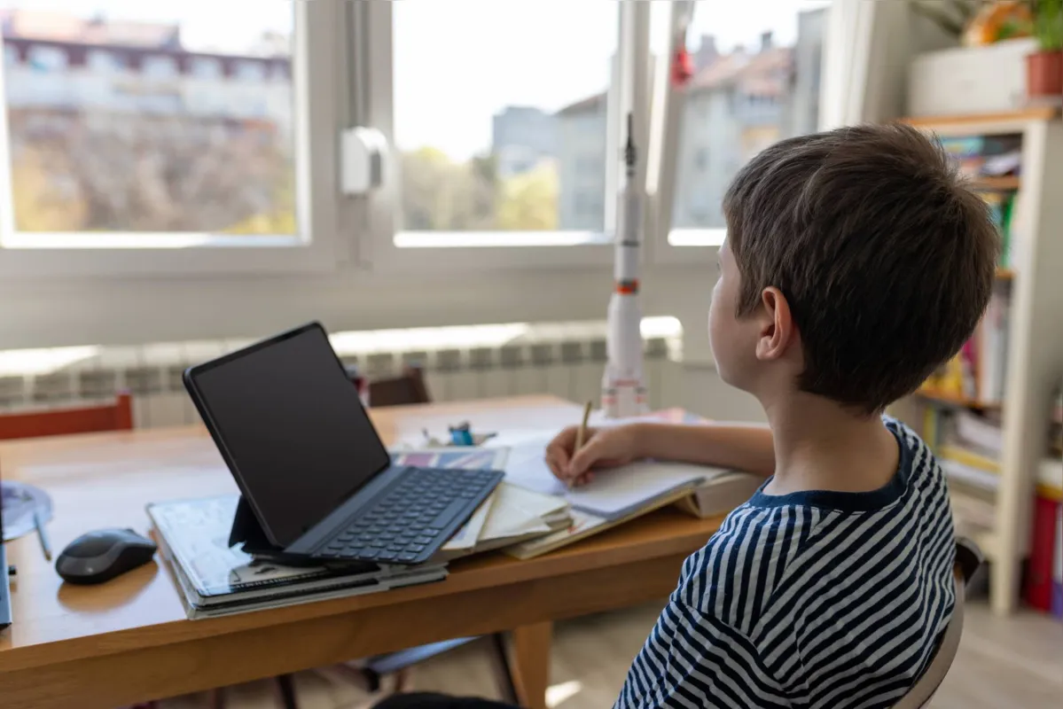 Elementary age boy sitting at the table at home, writing in his notebook and watching online lesson on laptop at time of Covid-19 outbreak