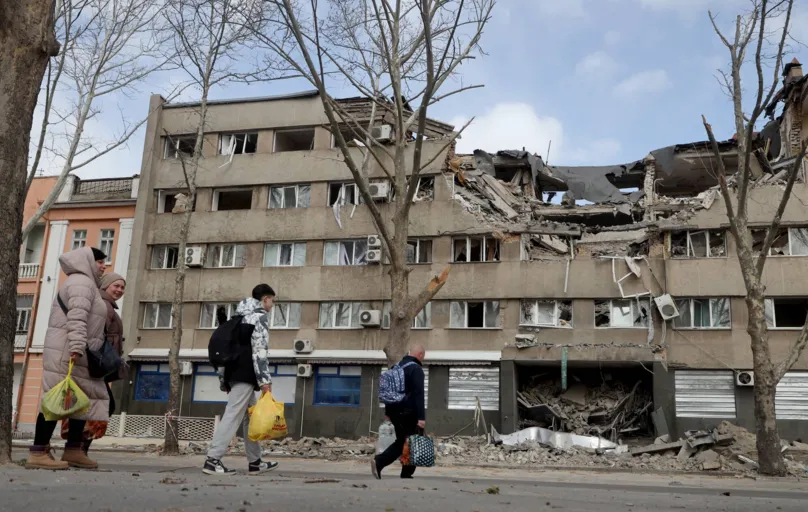 People walk past a damaged building in the Ukrainian city of Mykolaiv on March 27, 2022. - Over the weekend, the air raid sirens were no longer disturbing locals in Mykolaiv, who were increasingly venturing out on the streets. Most barely hurry when they hear them. (Photo by Oleksandr GIMANOV / AFP)