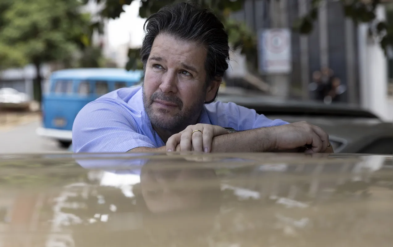Murilo Benicio debruçado sobre uma Caravan na cena em que sai da FOLHA interpretando o repórter Paulo Ubiratan